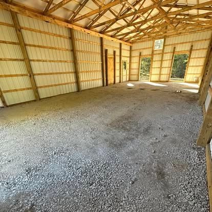 Empty wooden barn interior with gravel floor and exposed roof trusses, lit by sunlight from open doors and windows