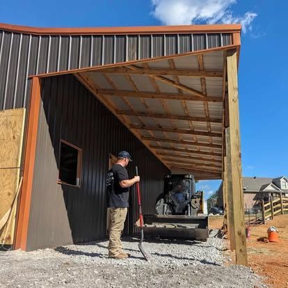 Worker standing beside a partially built metal garage with an open front and gravel ground.