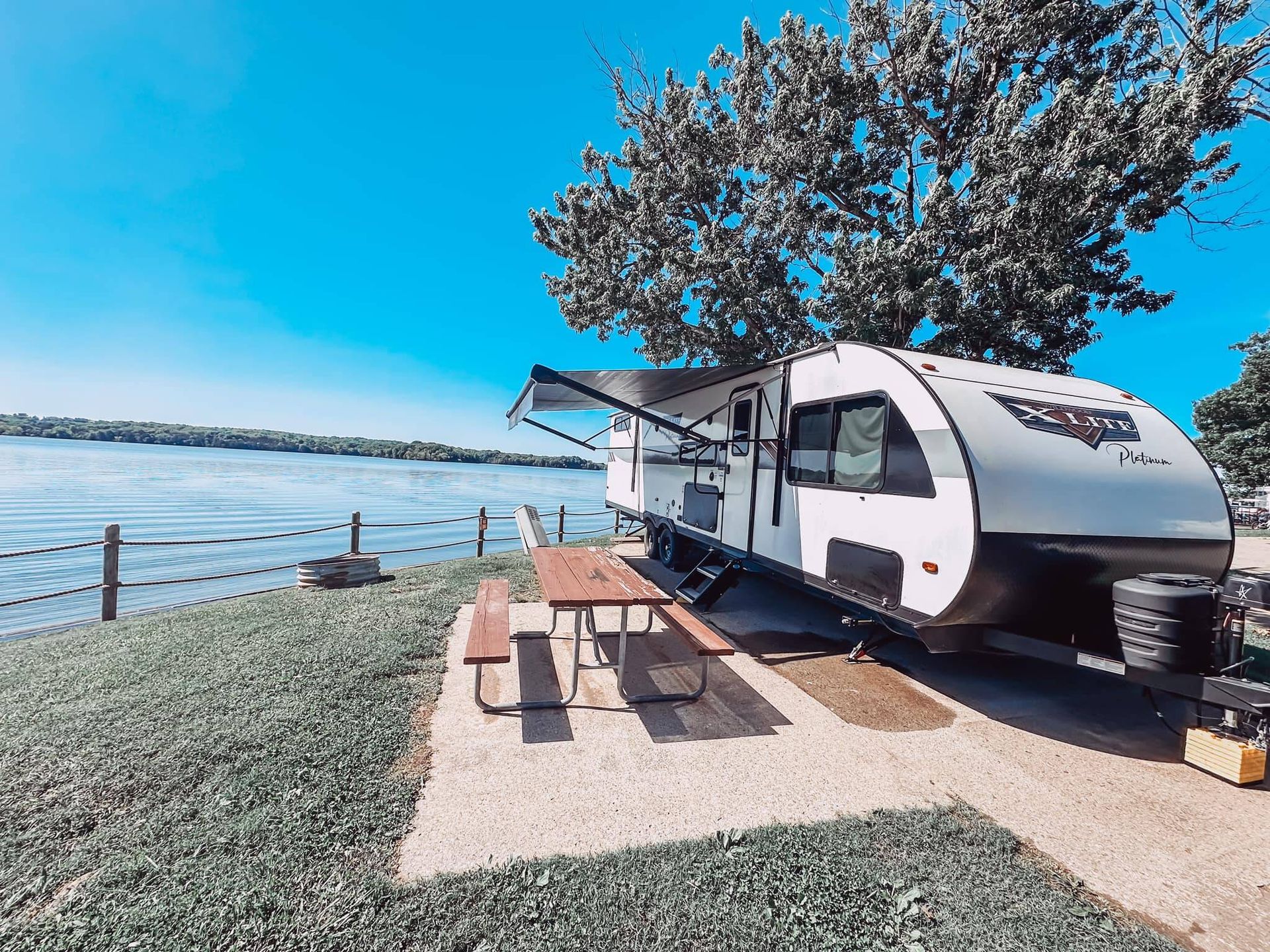 White camper trailer parked by a lake under a tree on a sunny day