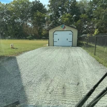 Gravel driveway leading to a small shed with a white garage door in a grassy yard.