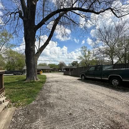 Gravel driveway beside a large tree, parked truck, and homes under a partly cloudy sky