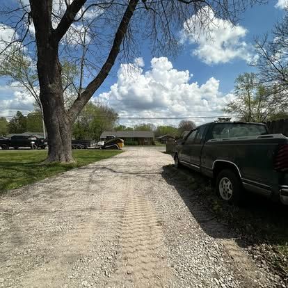 Gravel driveway beside a large tree and green pickup truck under a partly cloudy sky