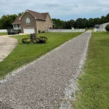 Gravel driveway beside a brick house with a white fence and green lawn.