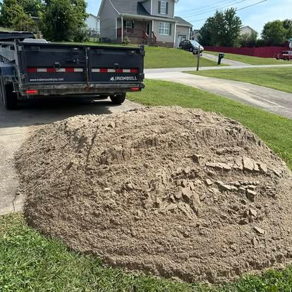 Large pile of dirt on a grassy lawn beside a driveway, with a dump trailer parked nearby.