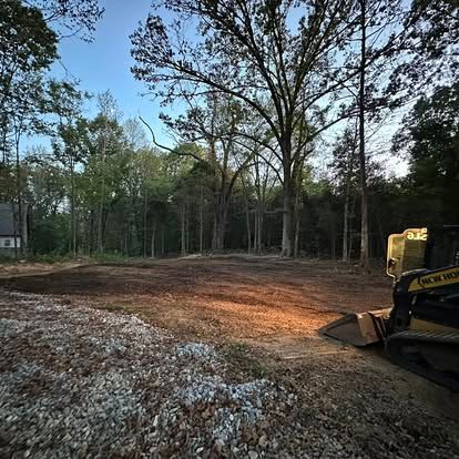 Excavator leveling a cleared dirt lot beside a wooded area at dusk