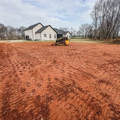 Small yellow skid steer leveling red dirt in front of a white house on a rural lot