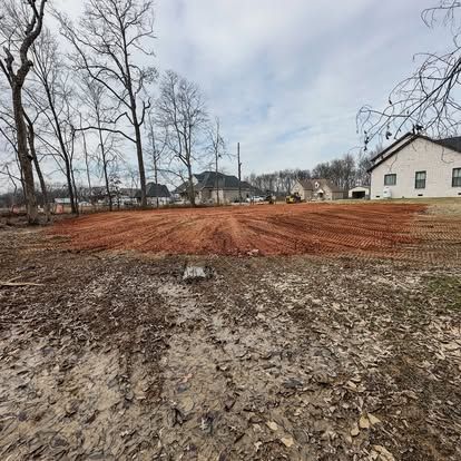 Bare muddy lot with a reddish dirt clearing, leafless trees, and a white house under a cloudy sky