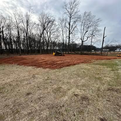 Small bulldozer grading reddish soil in a grassy field near leafless trees under a cloudy sky