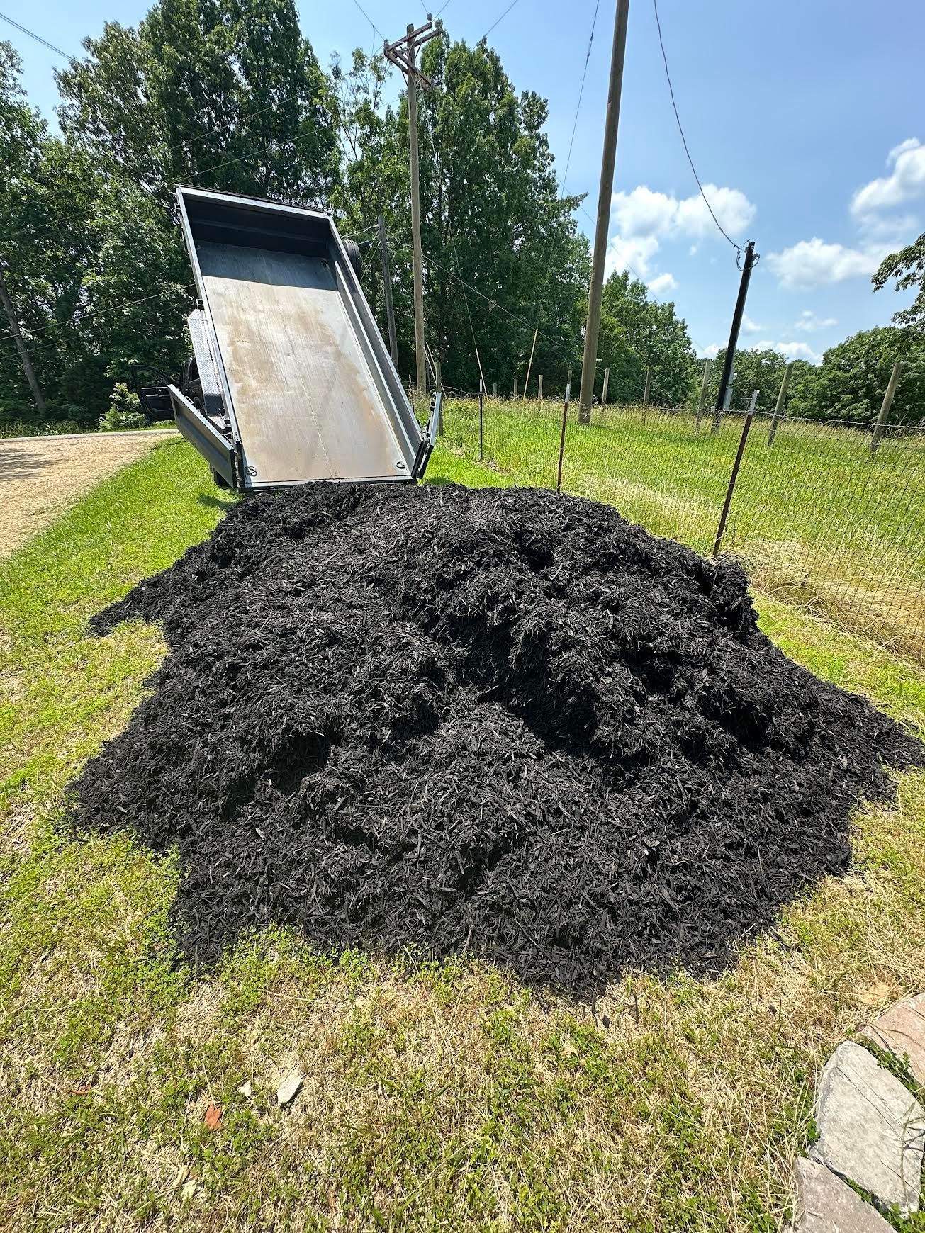Black mulch piled beside a dump trailer on a sunny grassy roadside