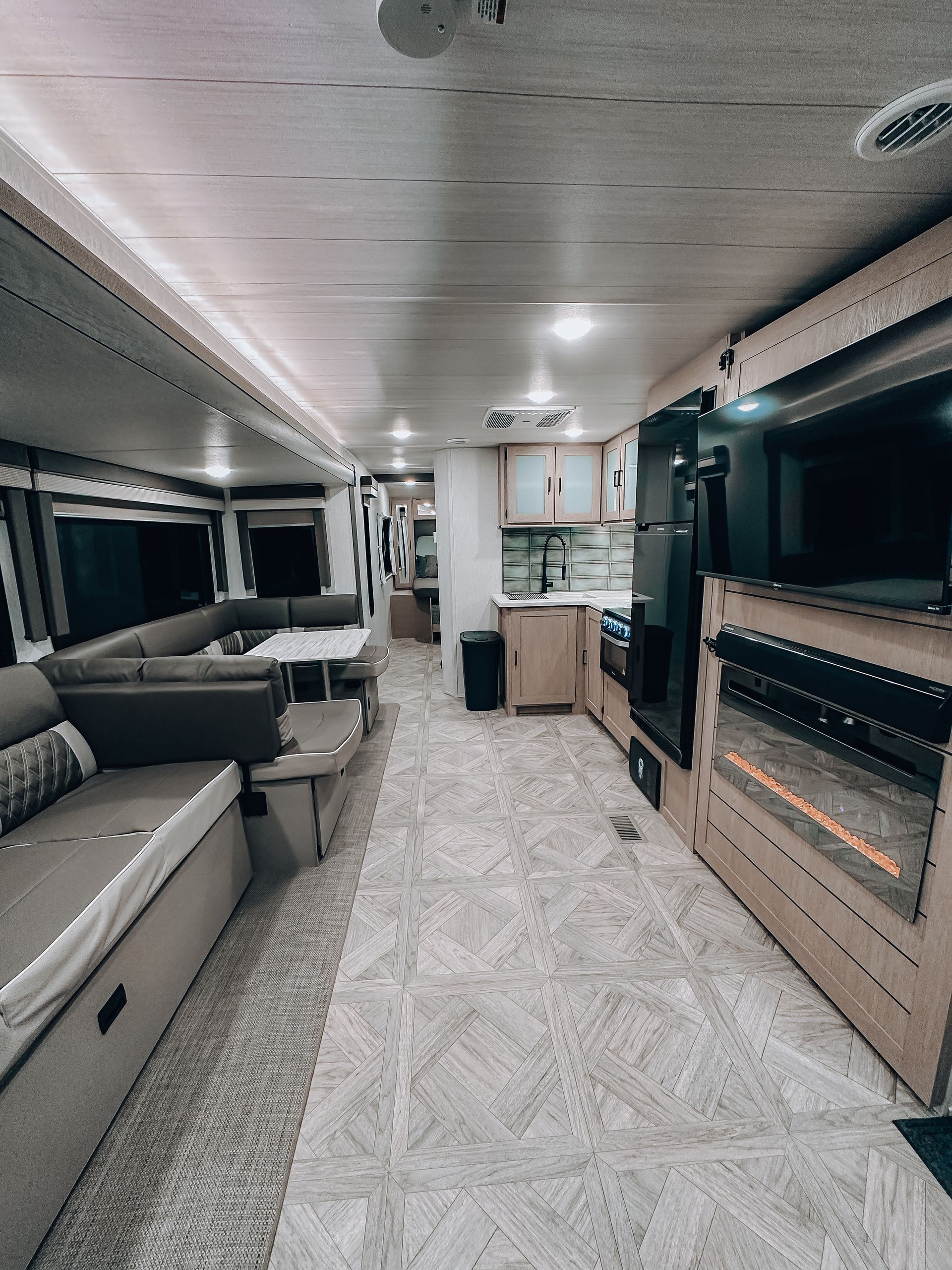 Modern RV interior with gray seating, a kitchen, and a large wall-mounted TV in a wood-paneled living area