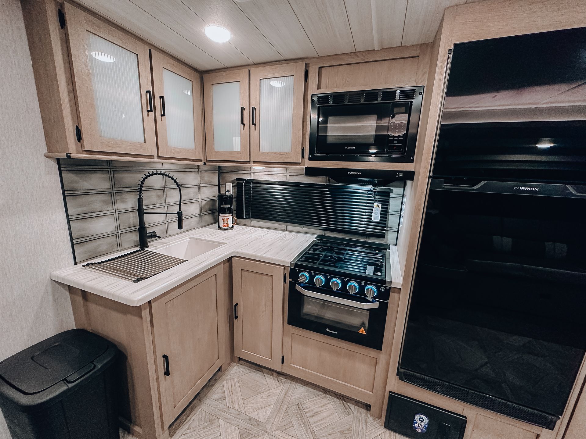 Compact RV kitchen with light wood cabinets, sink, stove, microwave, and black refrigerator