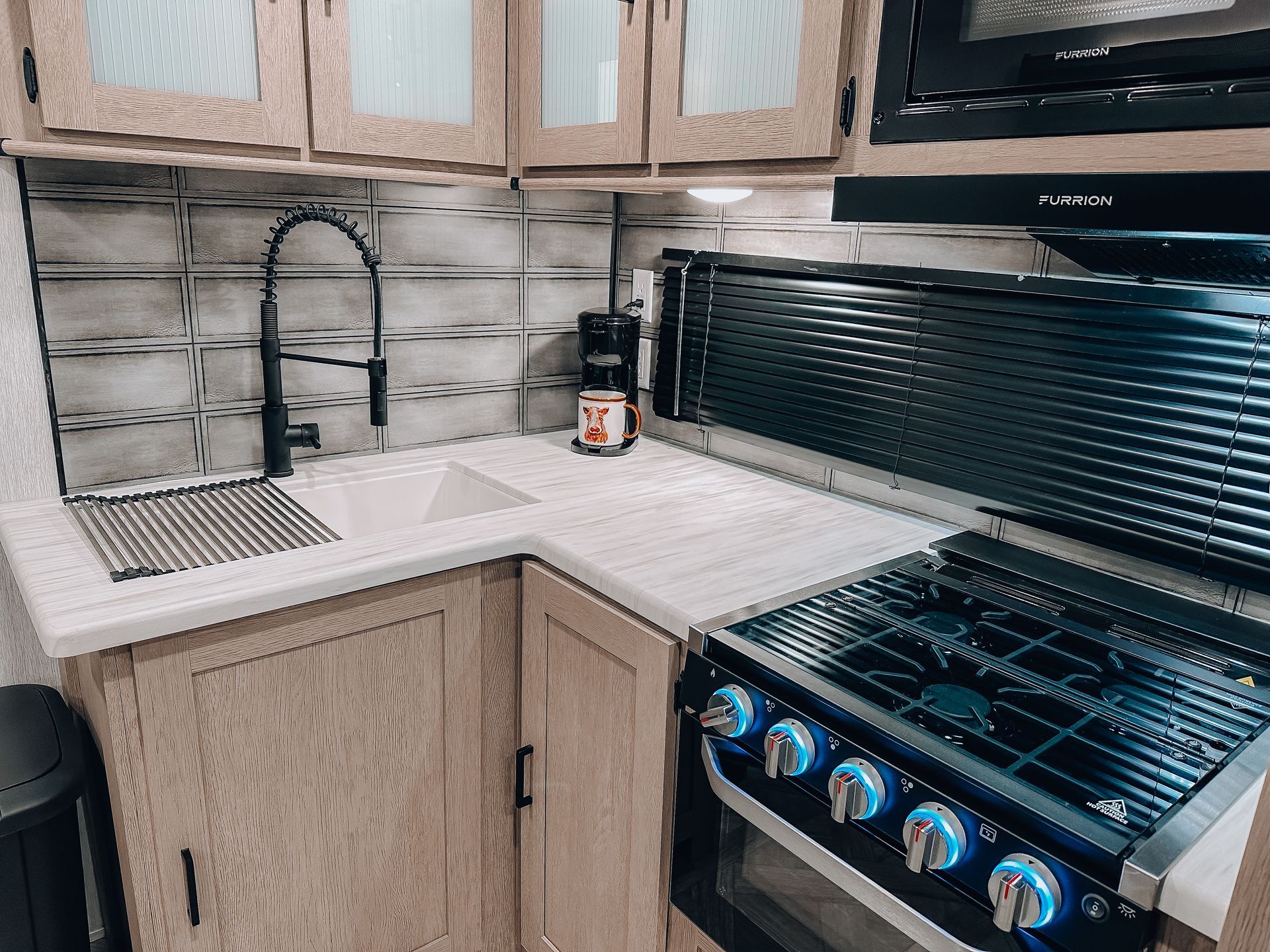 Compact kitchen corner with sink, dish rack, wood cabinets, and stainless gas stove under a window.