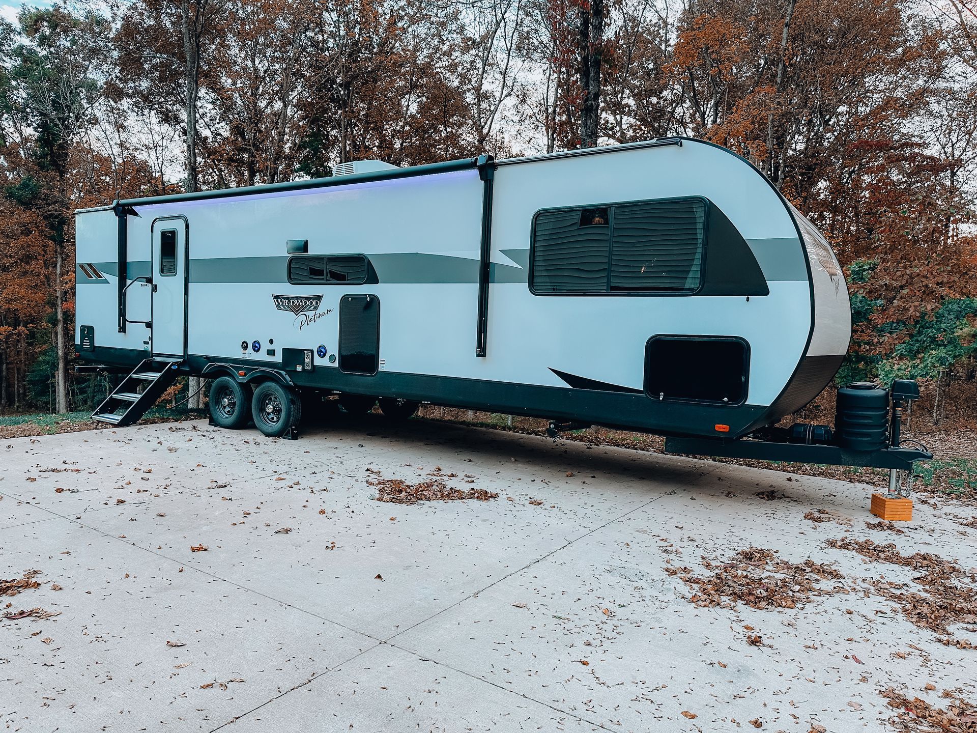 White travel trailer parked on a leaf-covered driveway beside autumn trees
