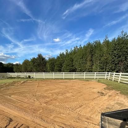 Riding arena with tan dirt, white fence, and trees under a blue sky