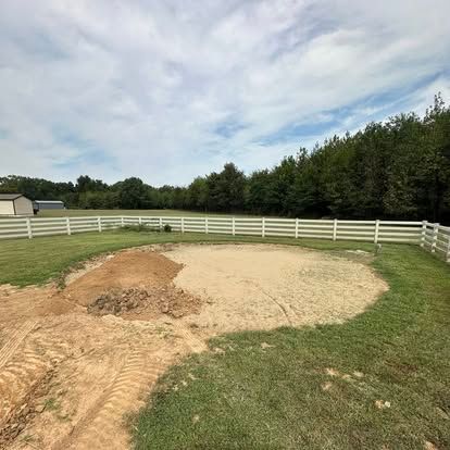 Gravel pad in a fenced grassy yard with a dirt pile and tree line under a cloudy sky