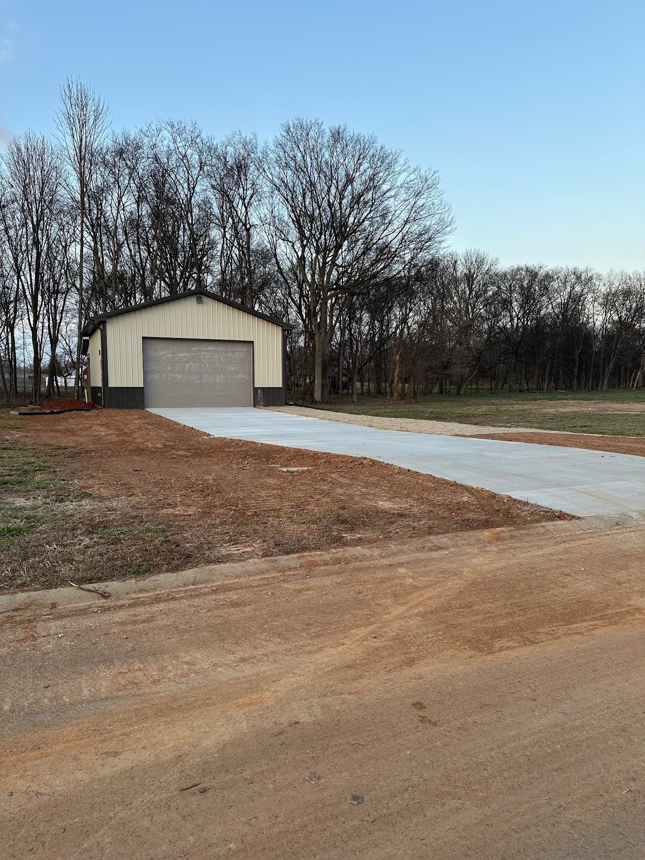 Small beige garage with a gray door at the end of a gravel driveway, bordered by bare trees.