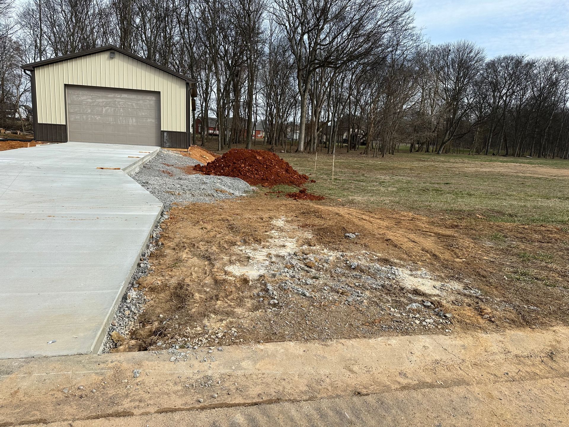 Concrete driveway leading to a beige garage, with gravel and dirt piles beside a grassy lot and bare trees