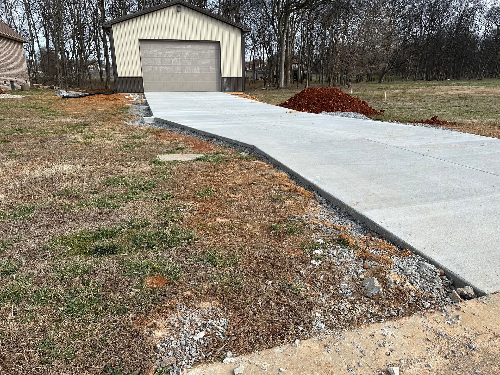 Concrete driveway leading to a beige garage in a rural yard with bare trees and patchy grass