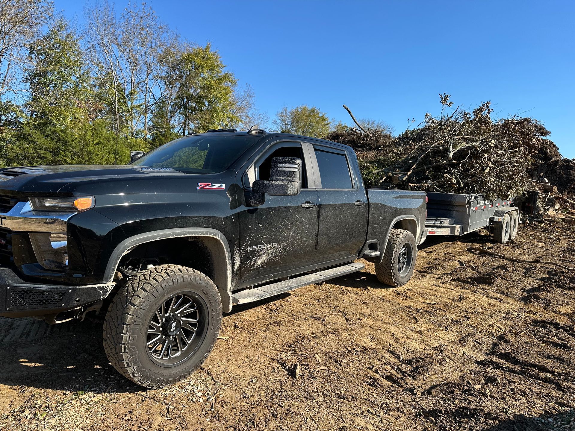 Black pickup truck hauling a trailer loaded with brush on a dirt lot under a blue sky