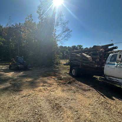 Sunny dirt lot with a log truck loaded with cut timber and a small loader near trees