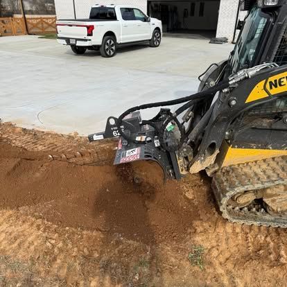 Excavator bucket digging brown soil near a concrete driveway, with a white pickup truck in the background