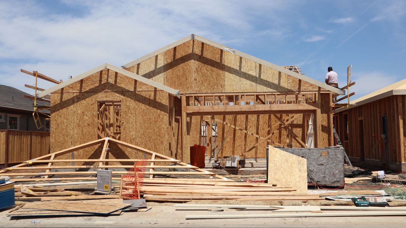Wood-framed house under construction with lumber piles, scaffolding, and workers on the roof and ground