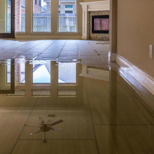 Interior flooded room, reflecting ceiling fan, windows, and fireplace. Beige walls, water-covered tile floor.