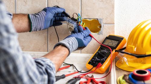 Electrician tests wiring at a wall outlet with a multimeter.