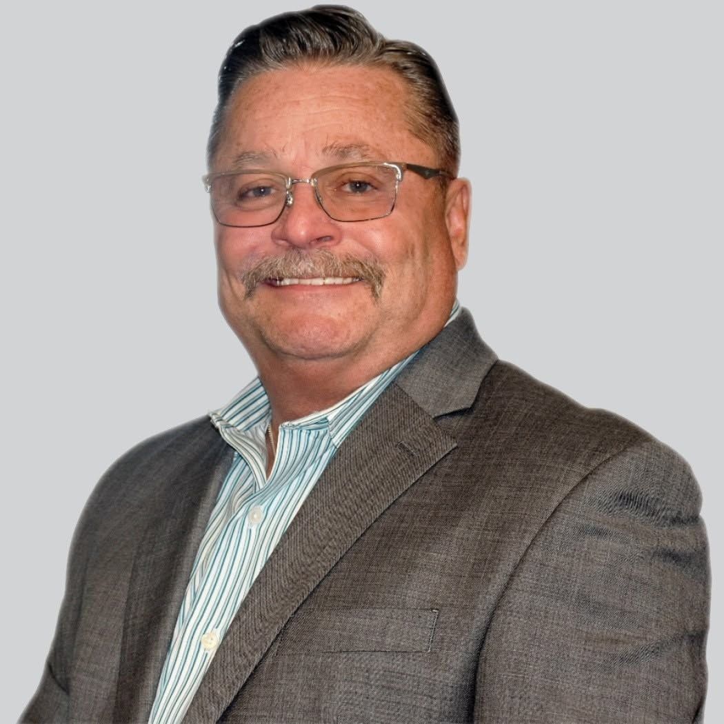 Smiling man in glasses wearing a gray blazer and striped shirt against a light gray background