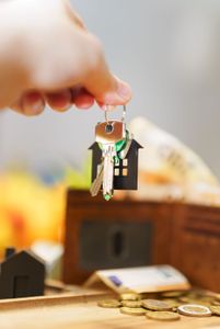 Hand placing a miniature house keychain above scattered coins and a small house model