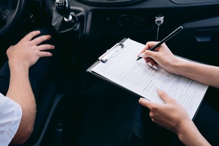 Hands in a car: one person holds a clipboard while another writes notes in the front seat.