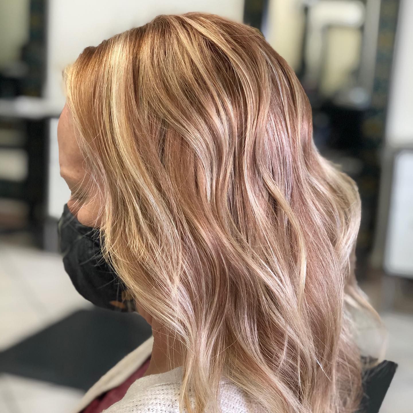 Side-view of wavy rose-gold blonde hair in a salon, with a black face mask and blurred background.