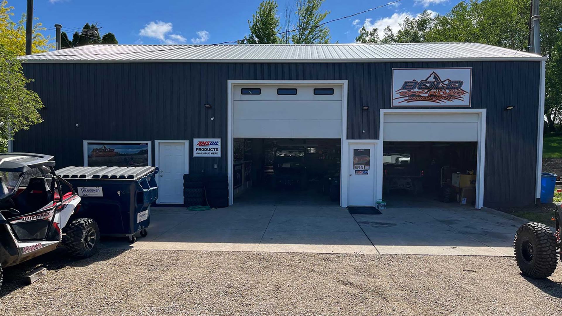 Blue auto repair shop with open garage doors, dumpsters, and a parked off-road vehicle outside