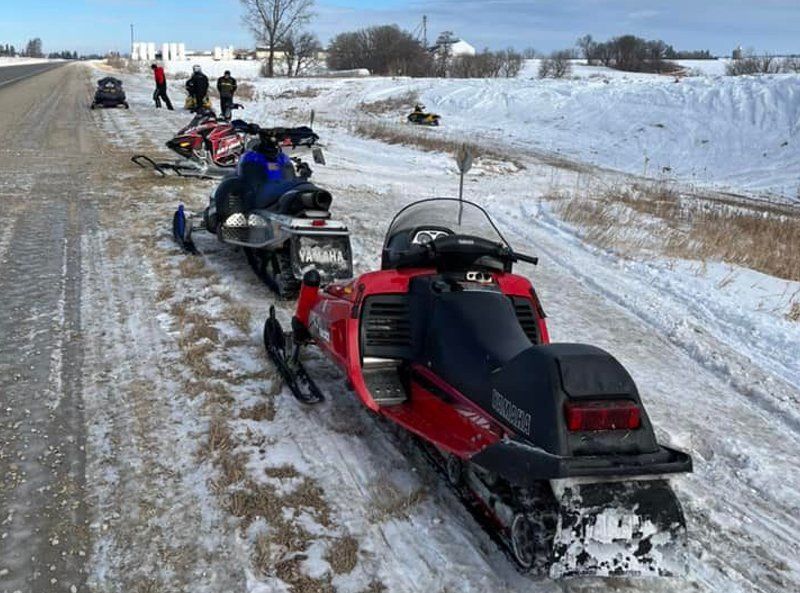 Snowmobiles lined up on a snowy roadside, with riders in the distance on a winter trail.