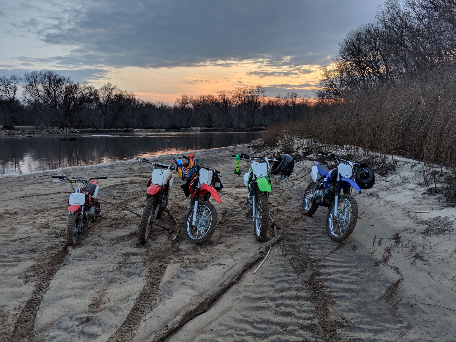 Five dirt bikes lined up on a sandy beach at sunset beside a calm lake.