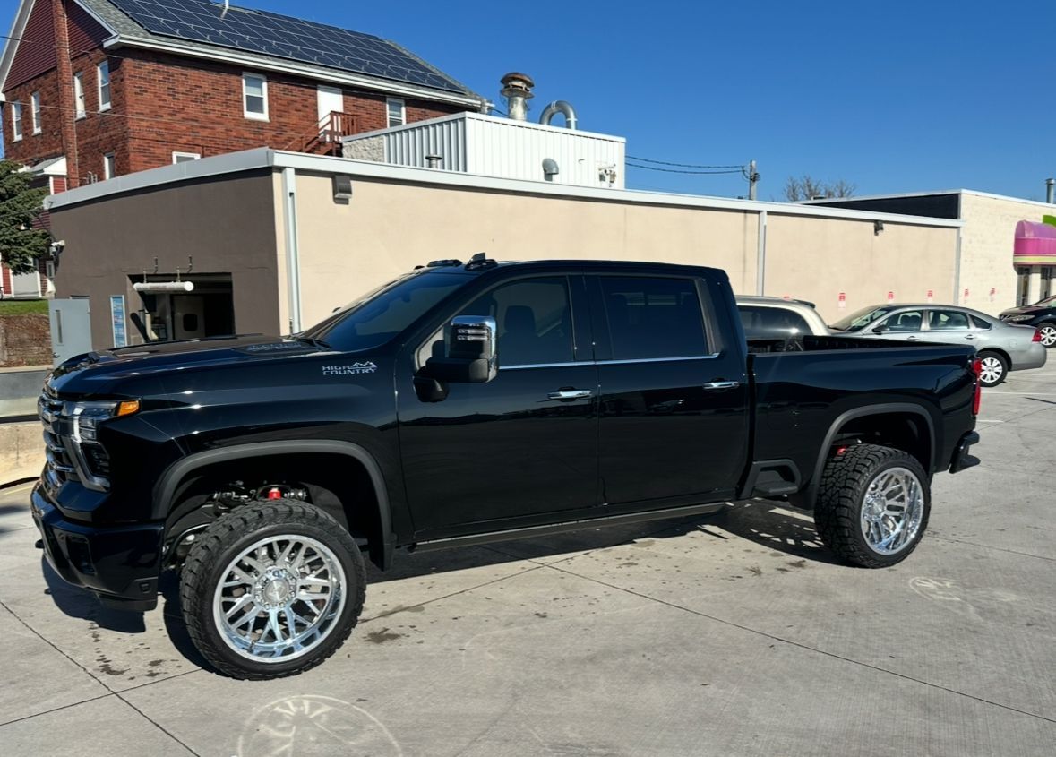 Black lifted pickup truck parked on a concrete lot beside a beige building and brick house.