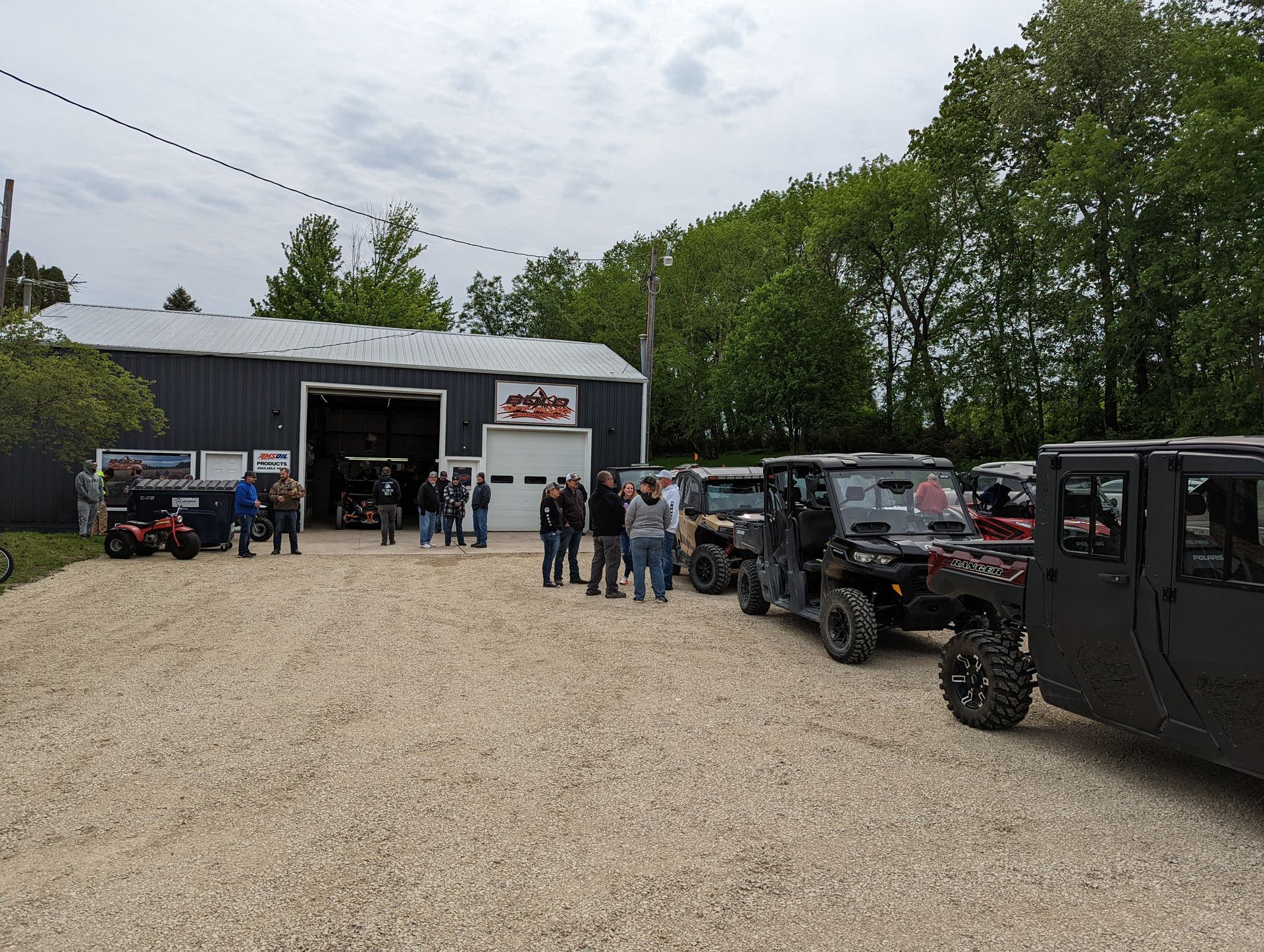 People gathered outside a garage with off-road vehicles parked on a gravel lot.