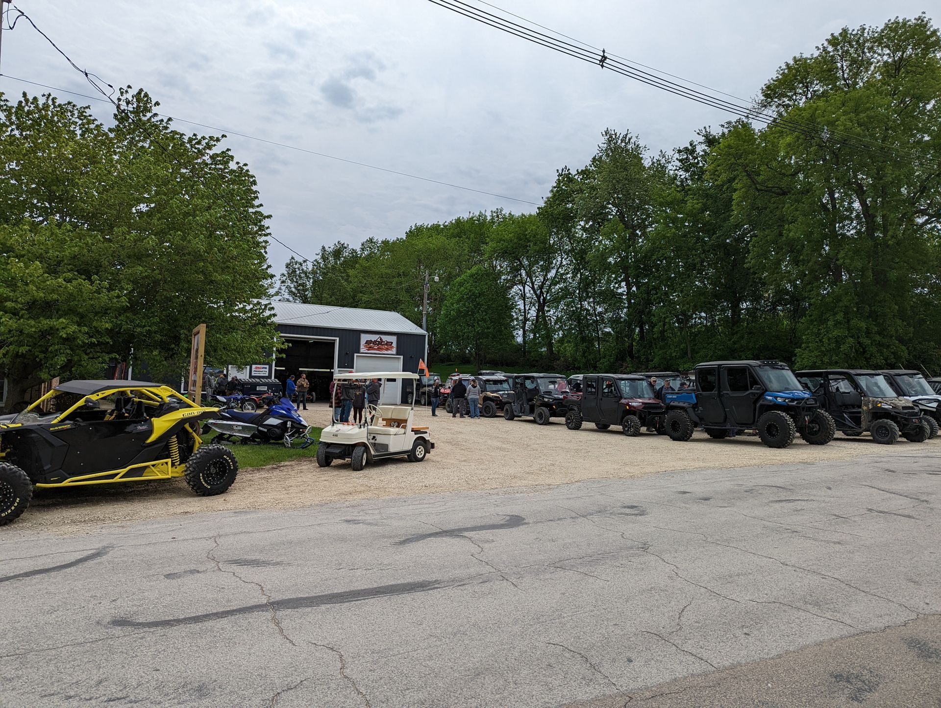 Off-road vehicles lined up on a gravel lot beside a small building and trees