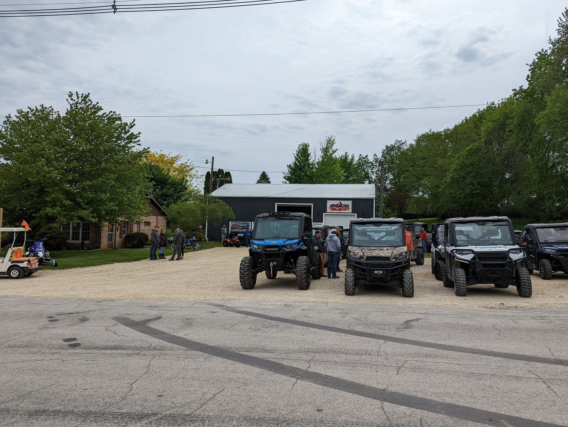 Four off-road vehicles parked on a gravel lot under a cloudy sky, with trees and a building behind them.