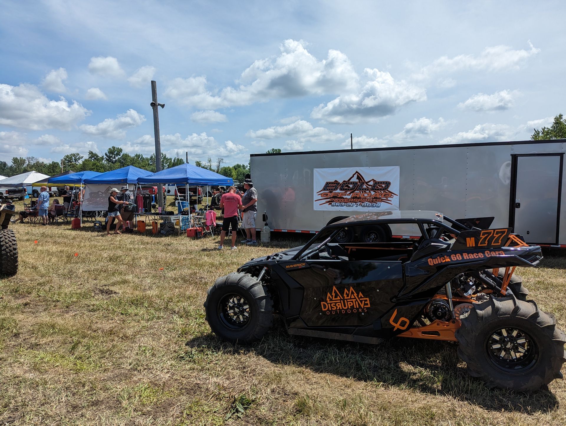 Black off-road buggy parked on grass at an outdoor event with tents and spectators under a cloudy sky