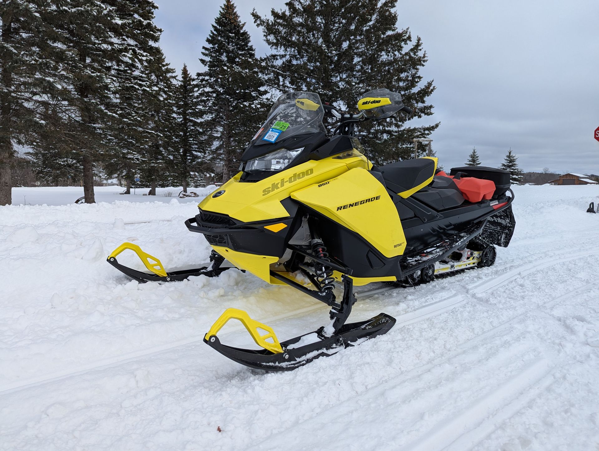Yellow snowmobile parked on snowy trail beside evergreen trees under a cloudy sky