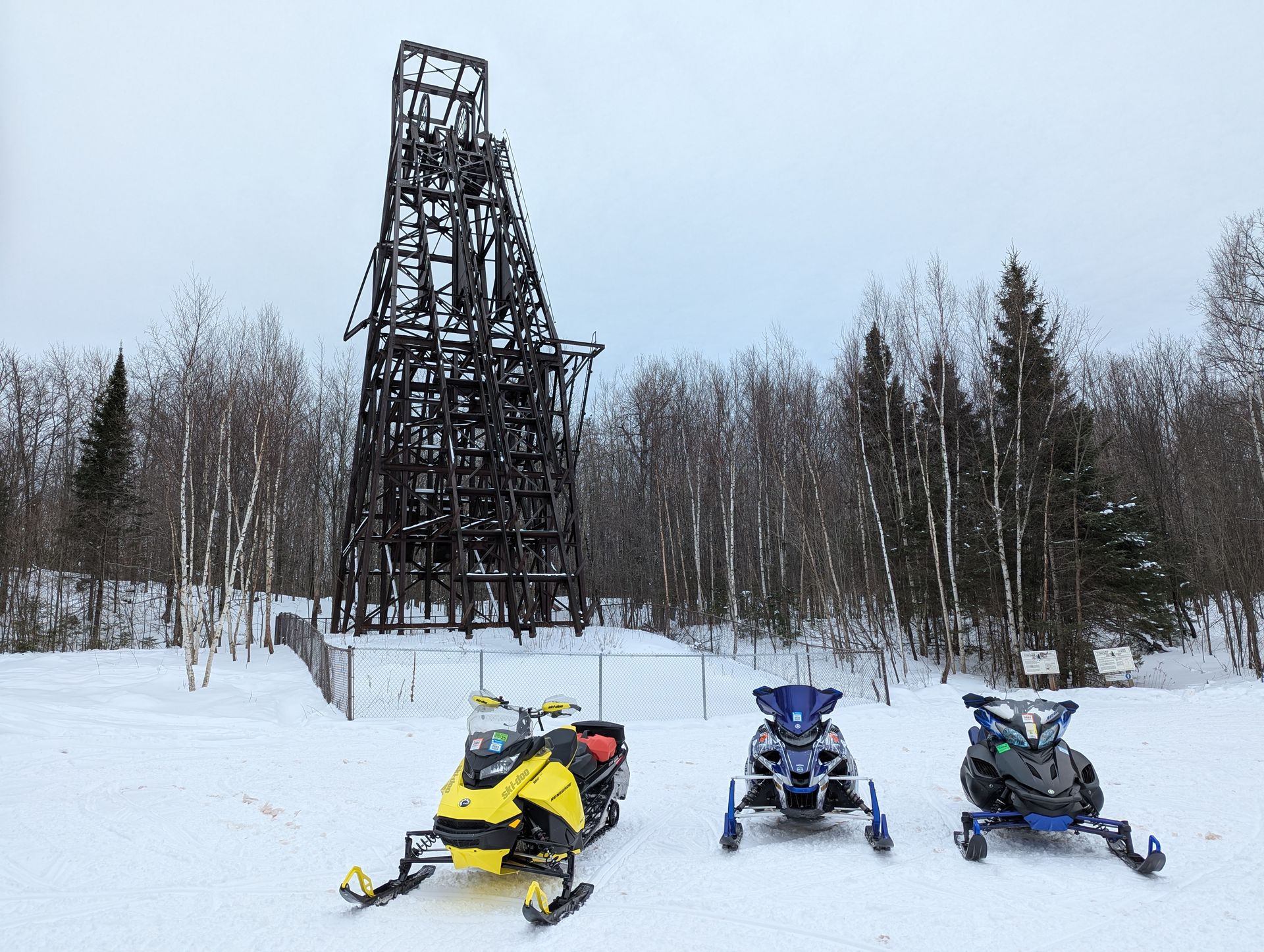 Three snowmobiles parked in snowy field before a tall dark tower and winter trees