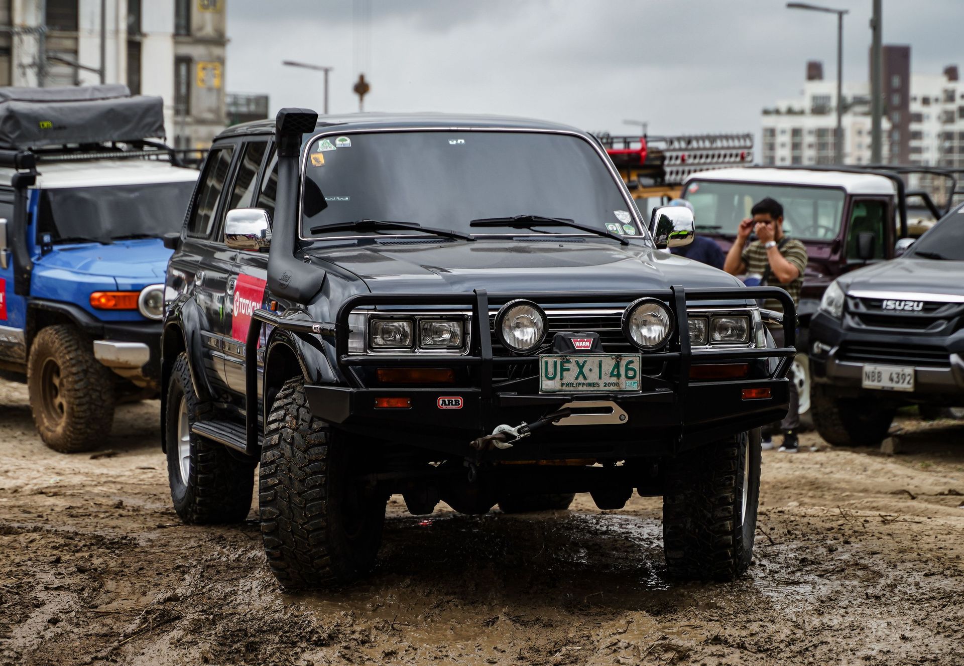 Black off-road truck in muddy lot, with other 4x4 vehicles and a driver taking a photo nearby.