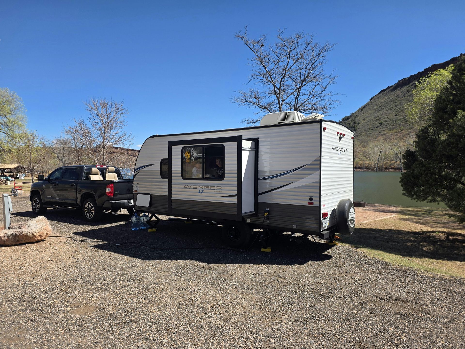 Silver camper trailer parked on gravel beside a black SUV under a clear blue sky