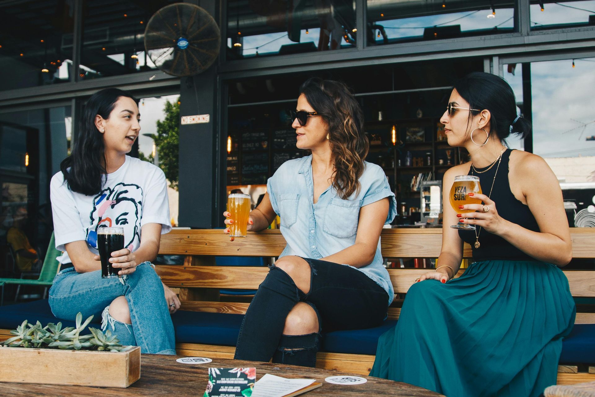 Three women seated outdoors at a table, drinking and talking. One wears a graphic tee, two wear sunglasses.