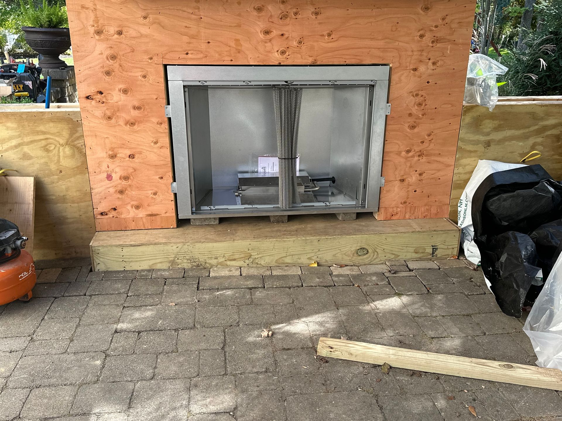 An unfinished outdoor fireplace framed in wood, centered on a paved patio with building materials nearby.