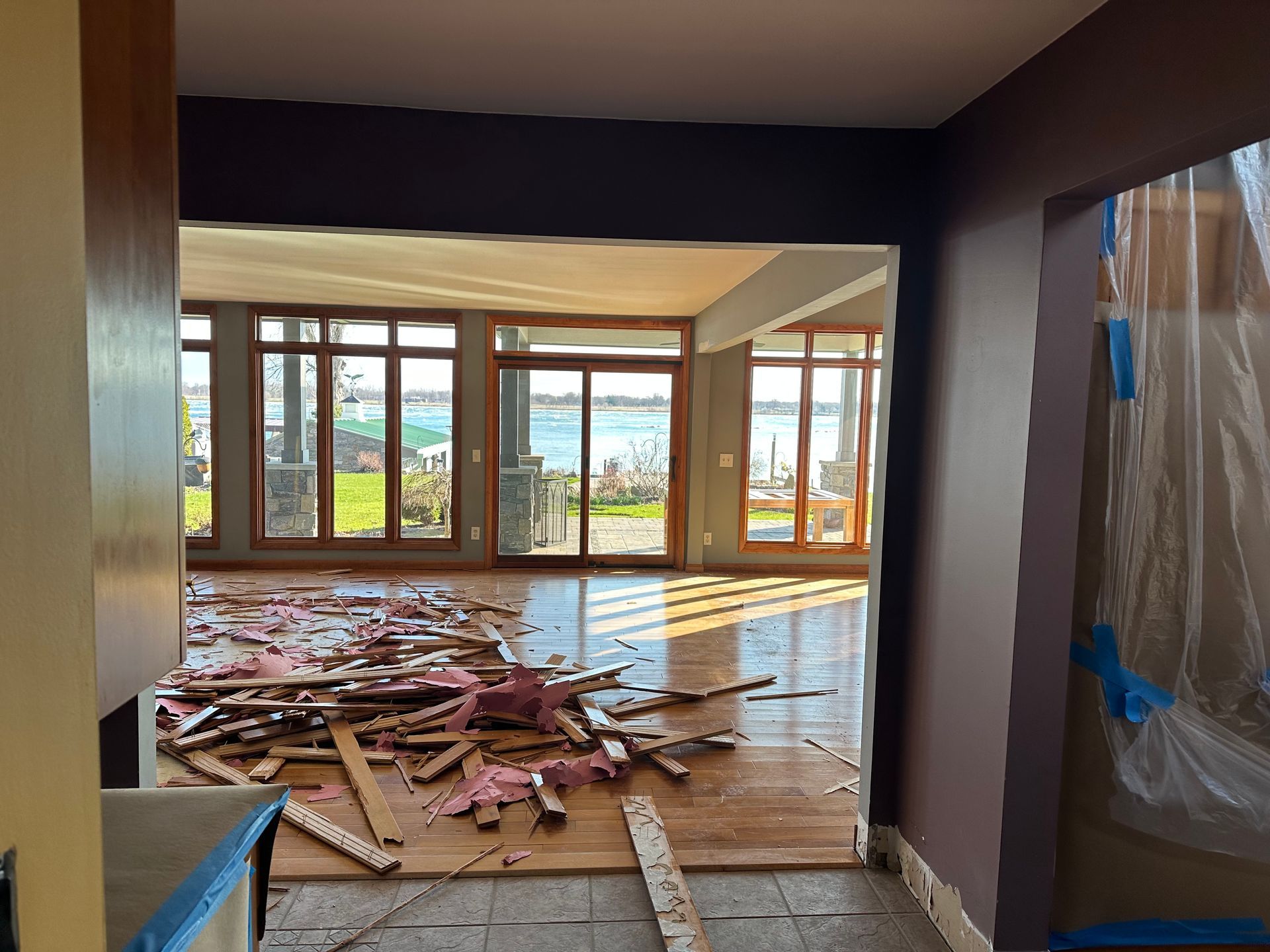 Renovation debris litters a room with wooden floors and large windows overlooking a body of water.