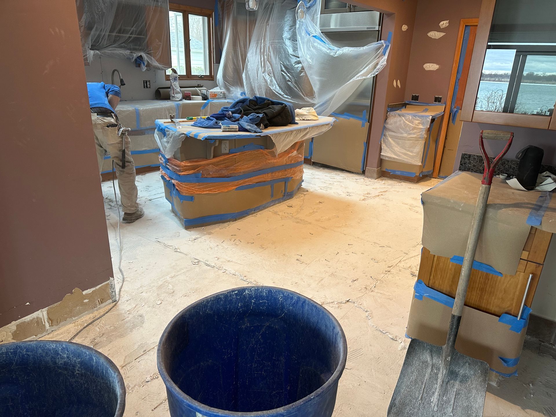 A room under renovation with covered countertops, drop cloths on the floor, a worker, and a shovel leaning against a crate.