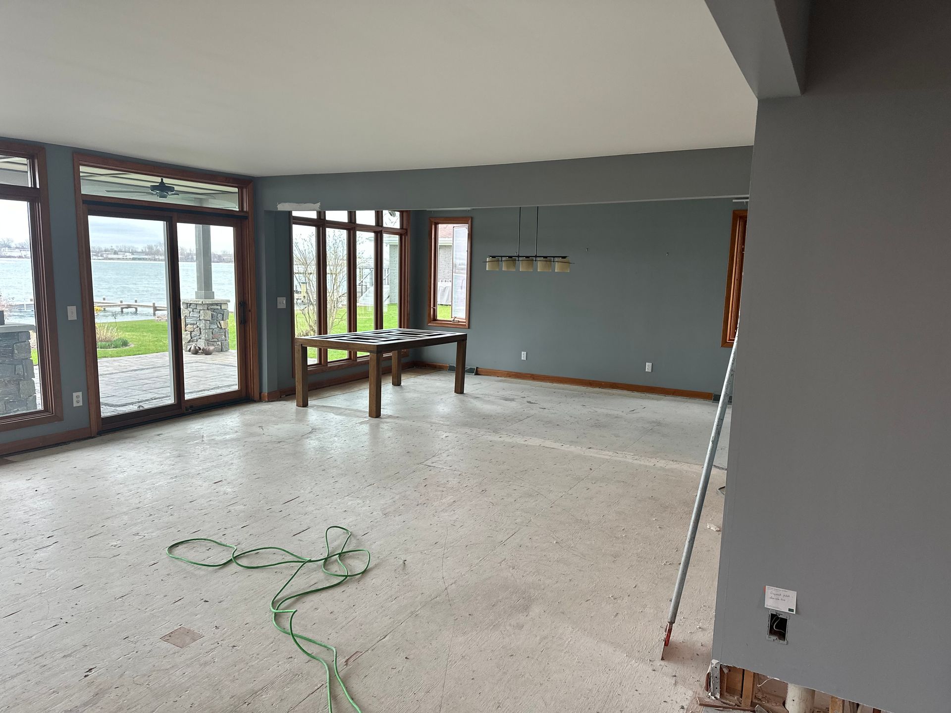 An interior room under renovation with gray walls, large windows overlooking water, a wooden table, and an exposed floor.