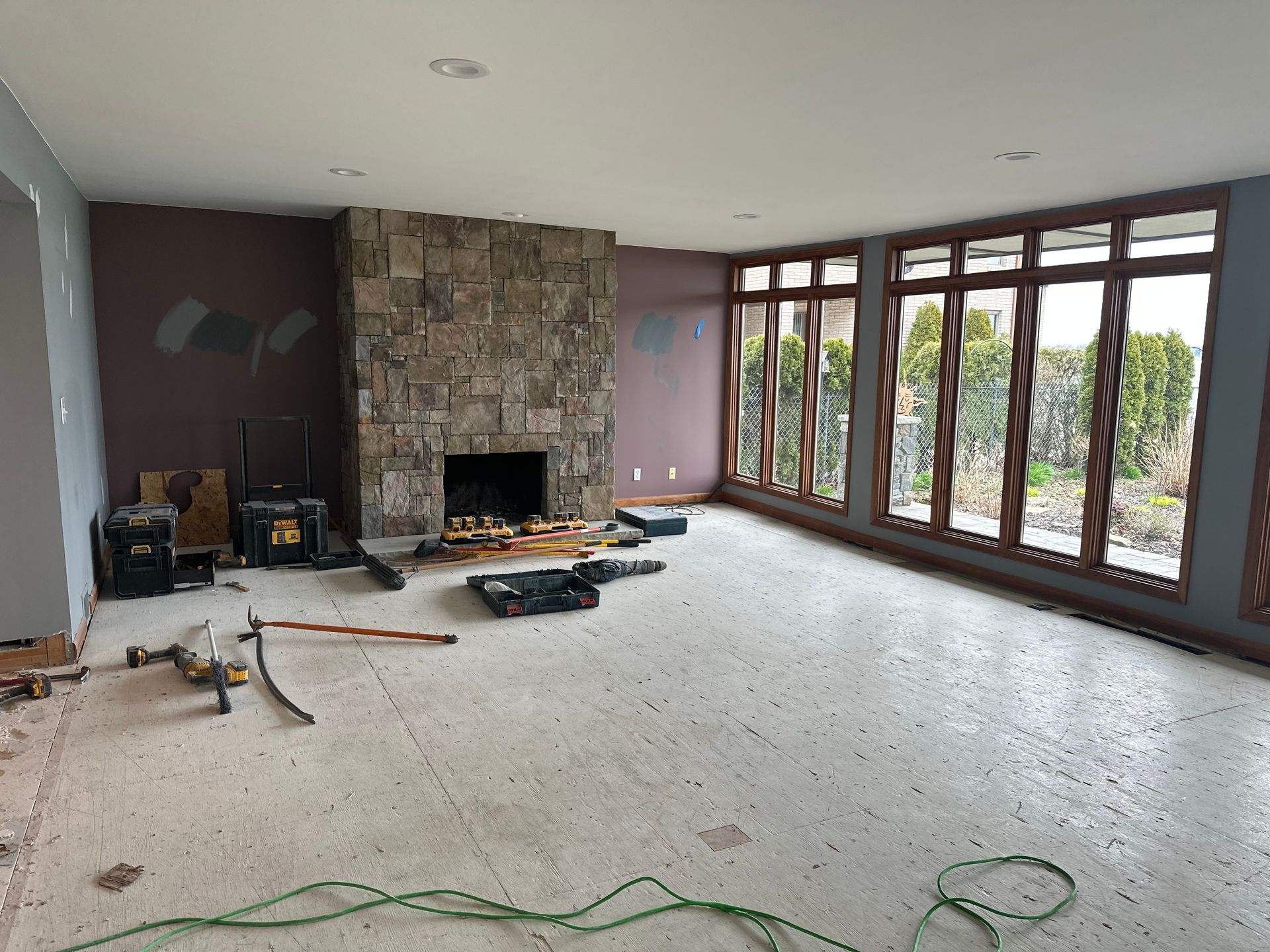 A room under renovation with a stone fireplace, dark walls, large windows, and construction tools on the concrete floor.